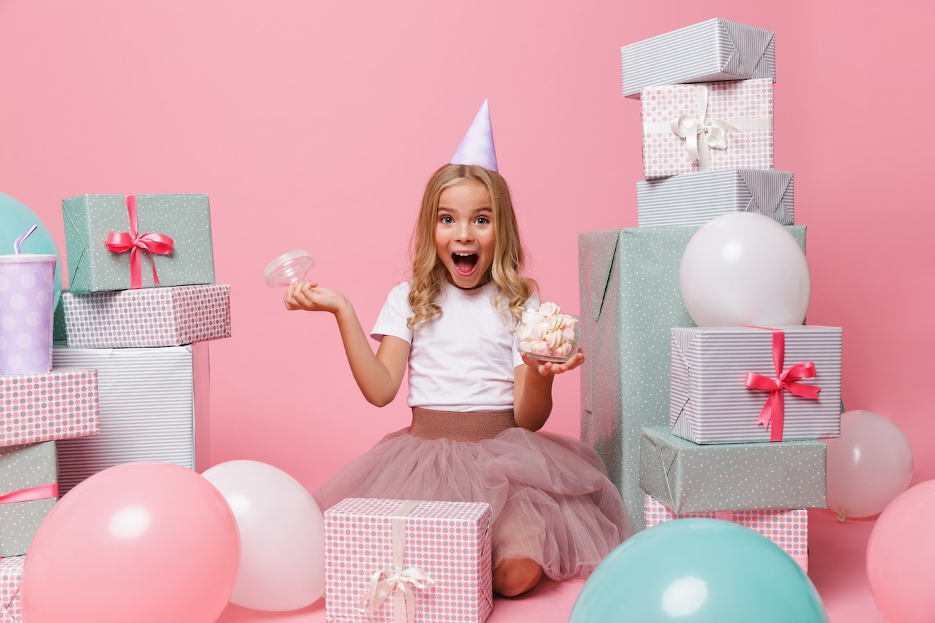Portrait of a joyful pretty little girl in a birthday hat celebrating while sitting in a pile of present boxes and holding jar of marshmallow isolated over pink background